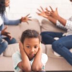 Unhappy child sitting in front of two parents fighting while sitting on a sofa - insights on can you lose parental rights for failing to co-parent from Kelly Jordan Family Law.